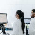 Dois pesquisadores usando jalecos e óculos analisam dados em um computador dentro de um laboratório moderno.