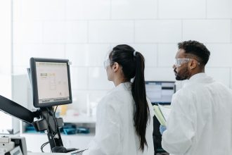 Dois pesquisadores usando jalecos e óculos analisam dados em um computador dentro de um laboratório moderno.