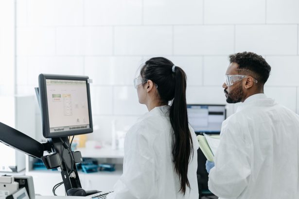 Dois pesquisadores usando jalecos e óculos analisam dados em um computador dentro de um laboratório moderno.