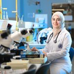 Pesquisadora trabalha em um laboratório diante de microscópios e livros de referência.