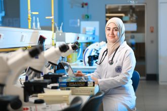 Pesquisadora trabalha em um laboratório diante de microscópios e livros de referência.