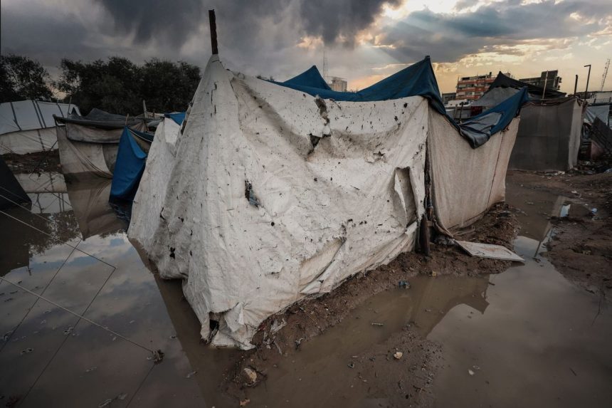 Abrigo de lona danificado em uma área alagada de um campo de refugiados em Gaza, sob céu carregado.