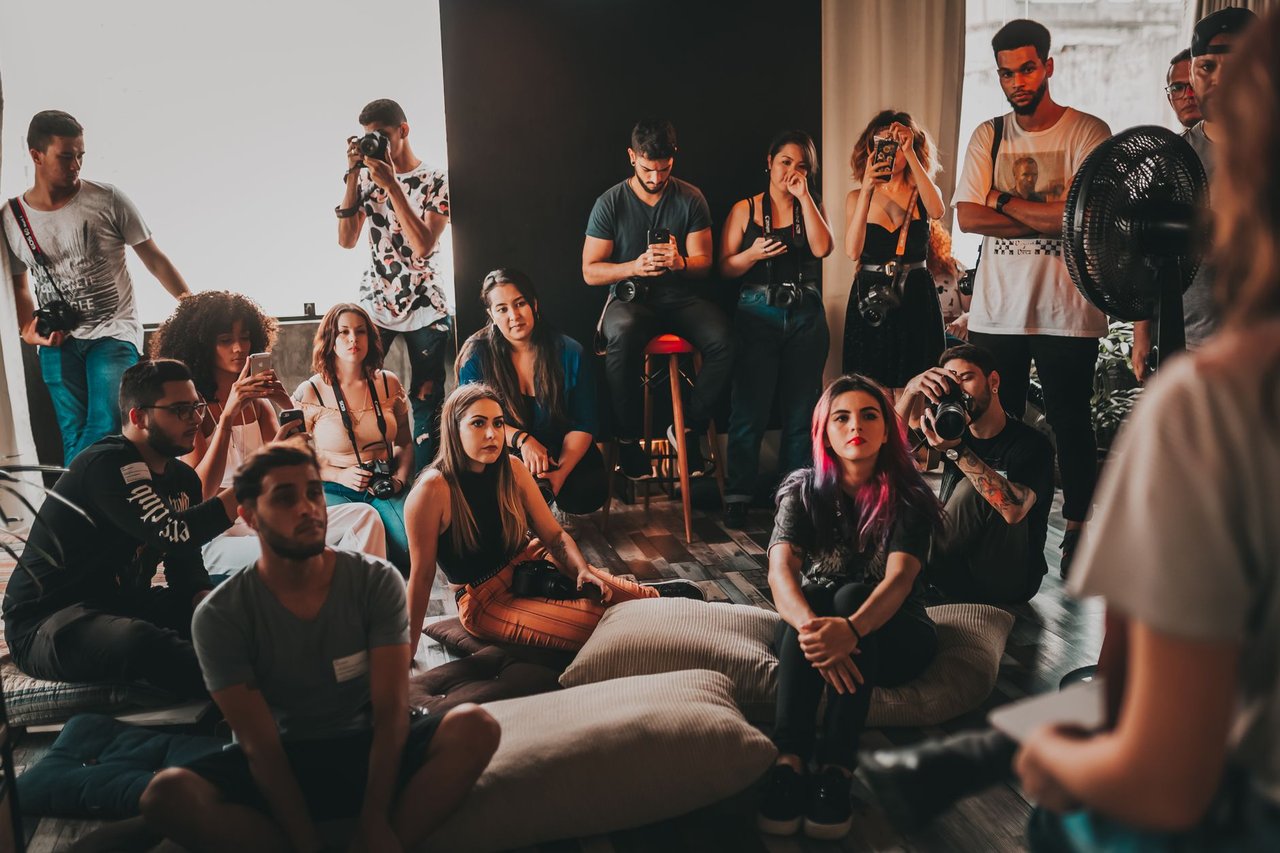 Grupo diverso de pessoas reunido em um espaço amplo, ouvindo atentamente uma mulher durante uma discussão.