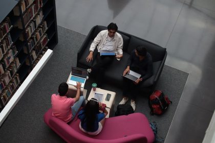 Grupo diverso de adultos colaborando em laptops em uma área de convivência de uma biblioteca universitária.