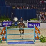 Cavaleiro conduz cavalo durante um salto sobre obstáculo em uma arena de competição equestre.