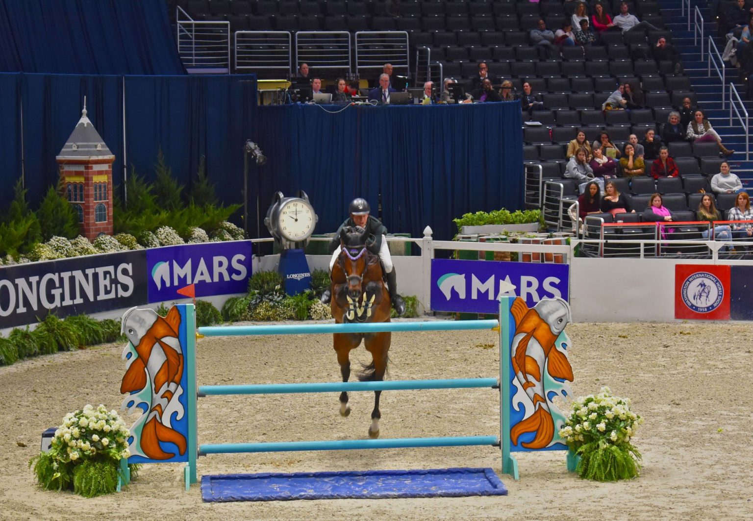 Cavaleiro conduz cavalo durante um salto sobre obstáculo em uma arena de competição equestre.