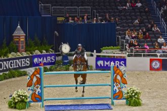 Cavaleiro conduz cavalo durante um salto sobre obstáculo em uma arena de competição equestre.