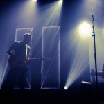 Músico toca guitarra diante de um microfone em um palco iluminado, enquanto um DJ aparece ao fundo.