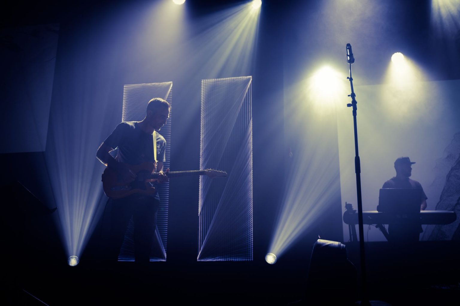 Músico toca guitarra diante de um microfone em um palco iluminado, enquanto um DJ aparece ao fundo.
