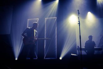 Músico toca guitarra diante de um microfone em um palco iluminado, enquanto um DJ aparece ao fundo.
