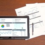 Computador, documentos e gráficos sobre uma mesa de trabalho, representando análise de informações e elaboração de relatório.